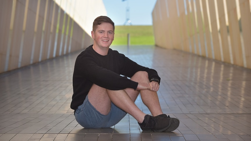 A young man is sitting on pavement, smiling at the camera with a blurred corridor behind him