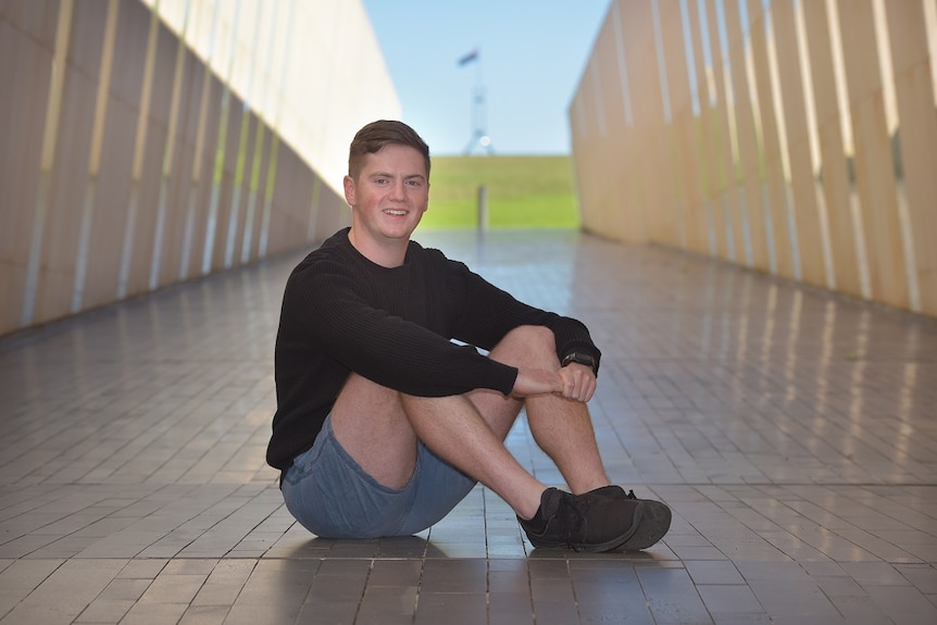 A young man is sitting on pavement, smiling at the camera with a blurred corridor behind him