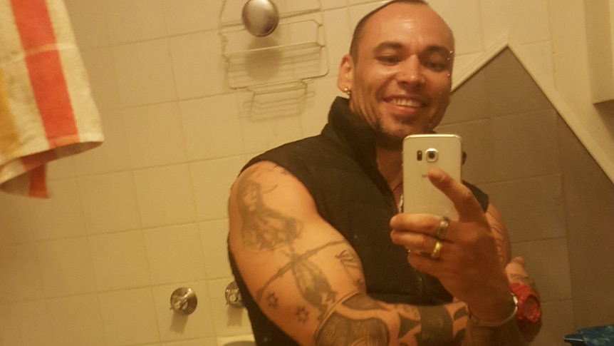 A young man with muscled arms full of tattoos smiles while taking a selfie in a bathroom.
