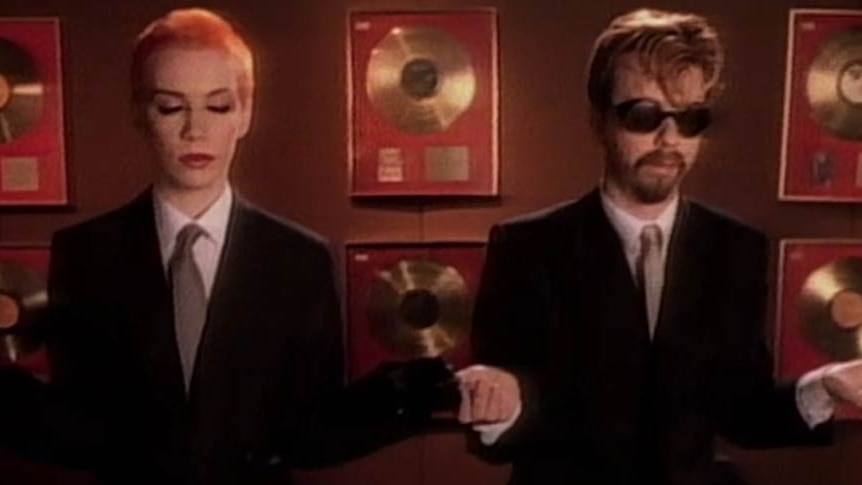 A man and woman meditate in front of a wall filled with gold records.