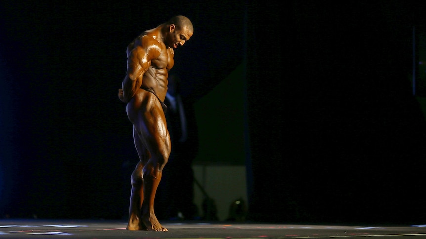 A black man with bulging, shiny muscles stands on a dark stage, illuminated by a spotlight. He looks down as he flexes