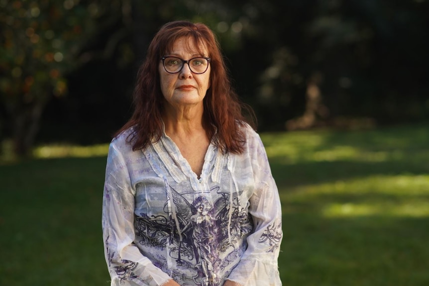 A bespectacled older woman with long, dark hair stands in a clearing.