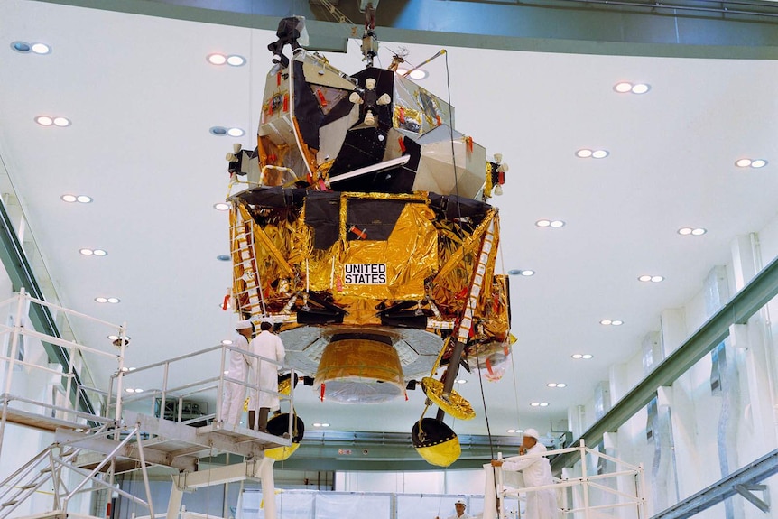 A lunar module hanging from a gantry in a factory overseen by workmen
