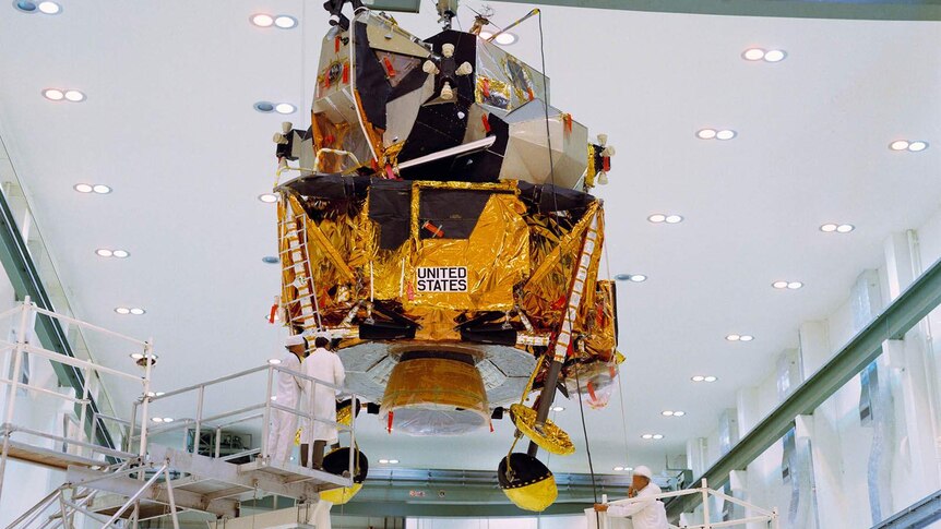 A lunar module hanging from a gantry in a factory overseen by workmen