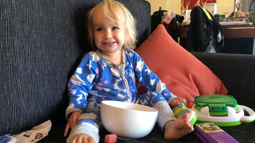 Blonde-haired toddler sitting on couch with bowl looking happy.