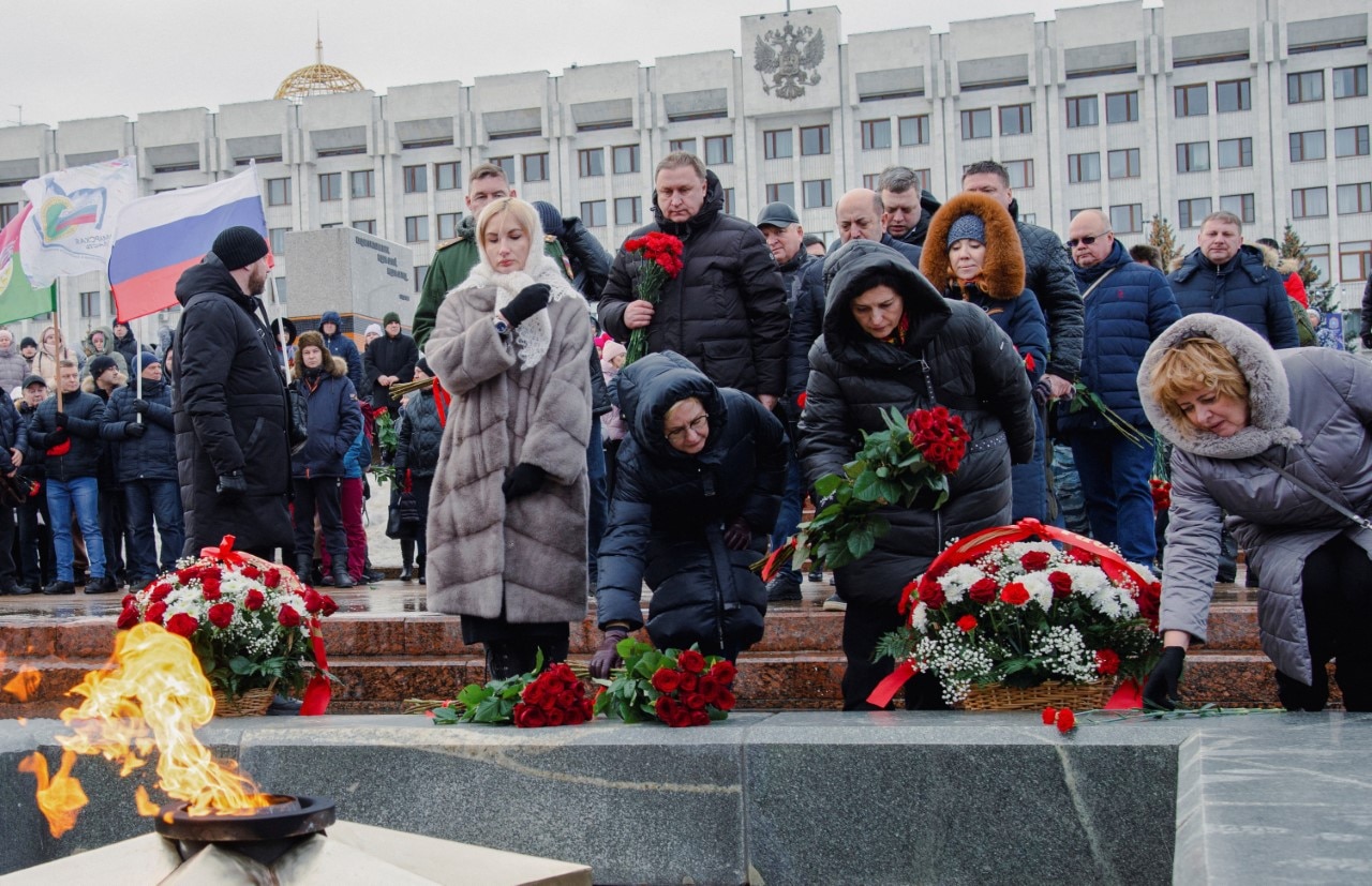 People lay wreaths and red flowers in a stone courtyard.