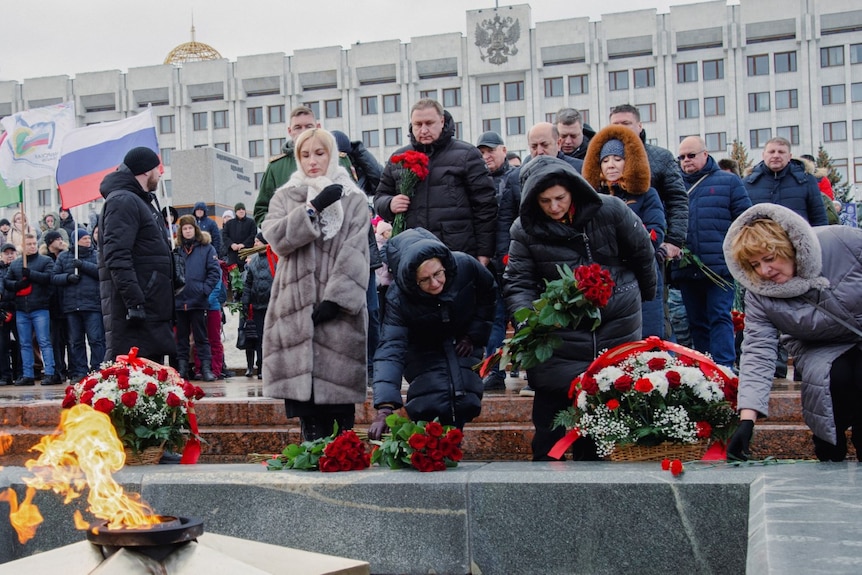 People lay wreaths and red flowers in a stone courtyard.