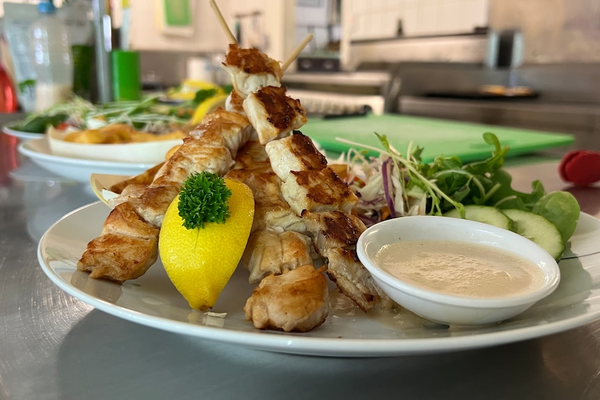 A close up photo of kebabs on a plate in front of dipping sauce and salad.