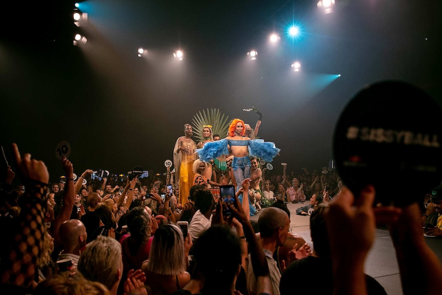 Bhenji Ra, dressed in blue, stands on the runway at Sissy Ball 2019, with members of the Slé voguing group standing behind her.