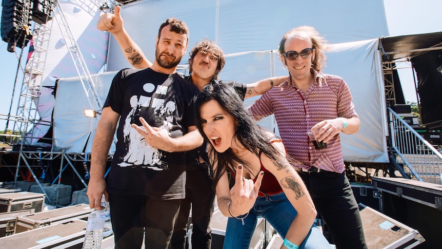 Four members of Magic Dirt backstage at a large concert. Adalita crouches down and give the devil horn rock hand signal