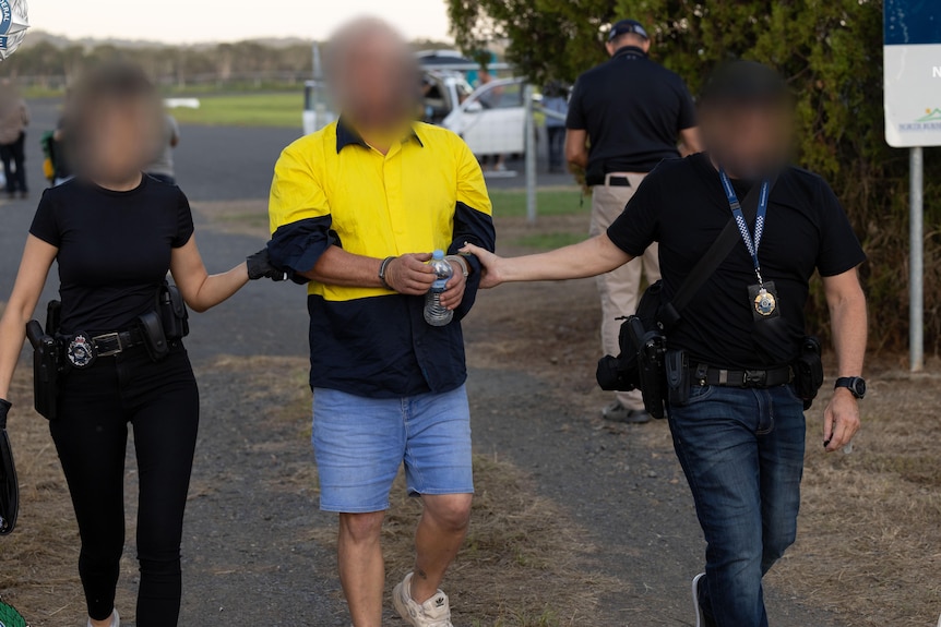 A man in a high-vis work shirt being led in handcuffs