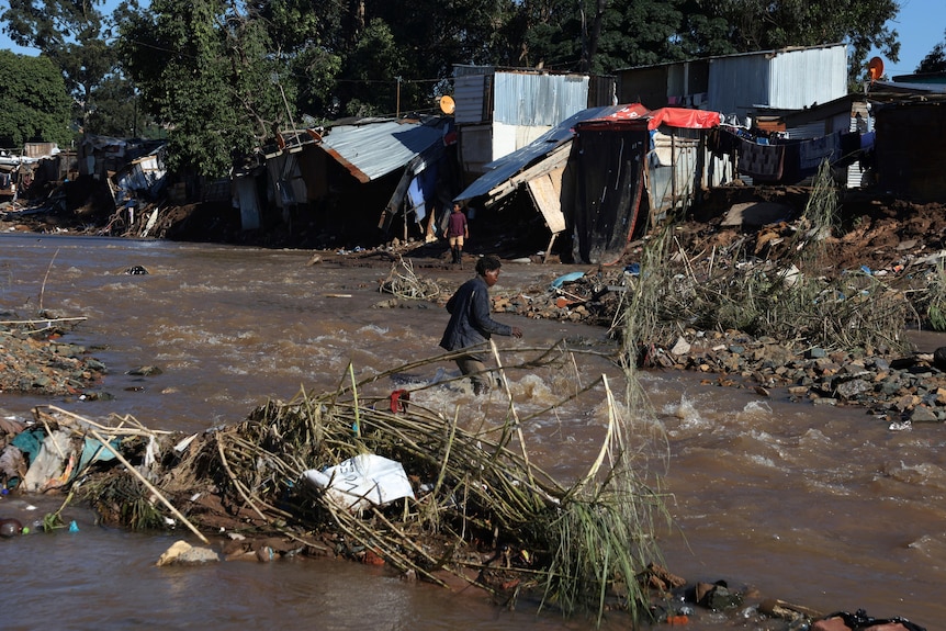 A man walks through a shallow river, amid rocks, broken tree branches and rubbish, near delapidated sheds.