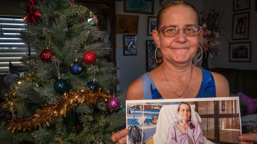 Woman standing in front of a Christmas tree holding a photo of herself in hospital with tubes in her neck