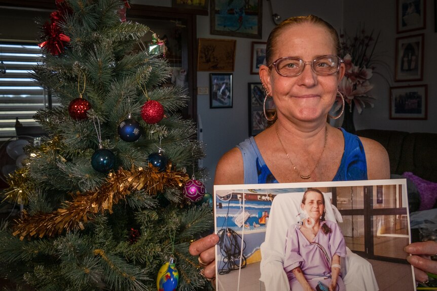 Woman standing in front of a Christmas tree holding a photo of herself in hospital with tubes in her neck