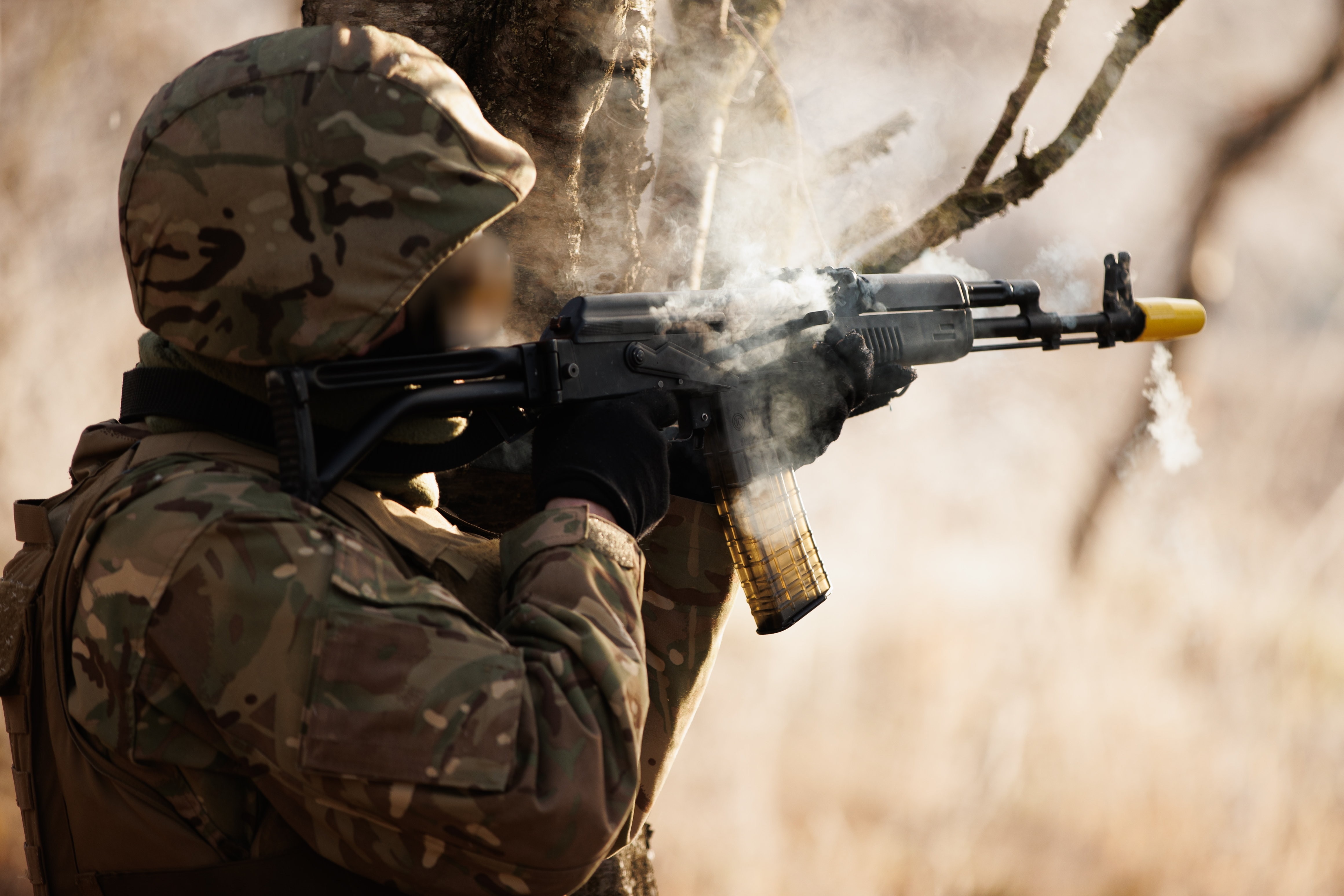 A soldier leaning up against a tree in khaki firing an assault weapon