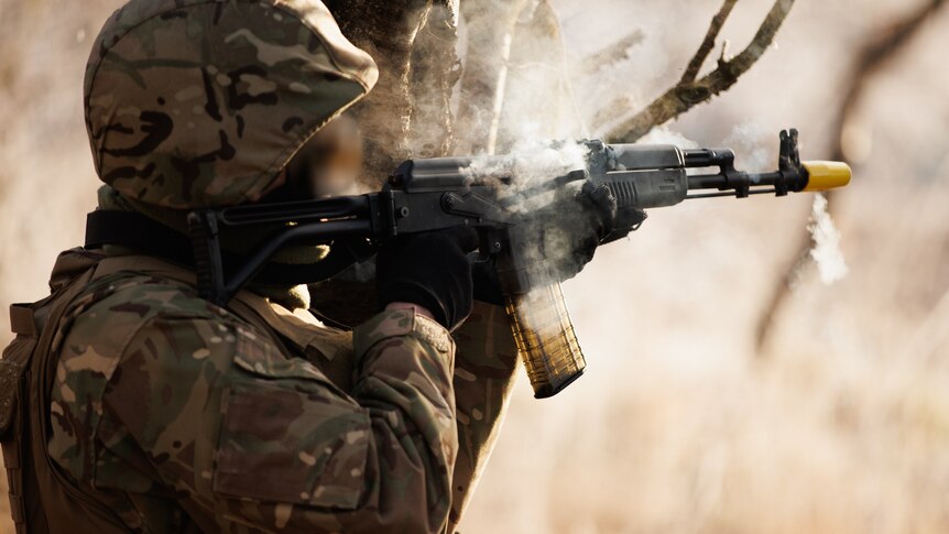 A soldier leaning up against a tree in khaki firing an assault weapon