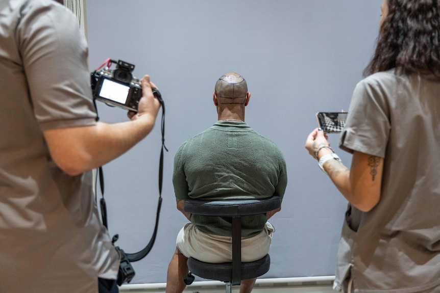 A man with shaved head and lines drawn around it sits in a chair, his back to two people with cameras