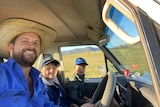 A man in a hat and a blue shirt takes a selfie at the wheel of a stopped car, with his two boys beside him.