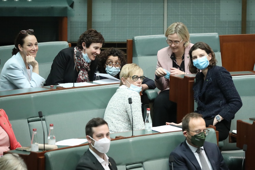 Six women gather at the end of a row of seats in the House of Representatives talking and smiling.