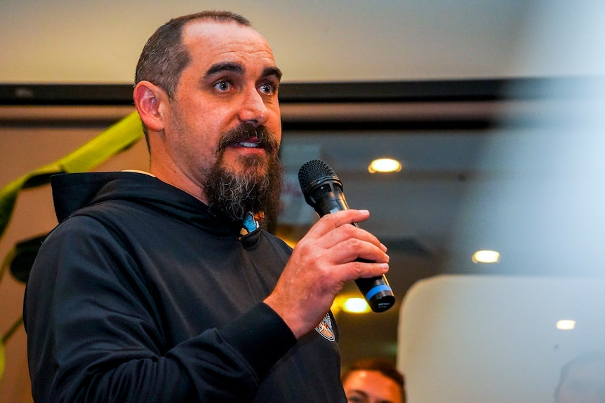 Rueben Berg speaks into a microphone as he stands at an indoor community event.