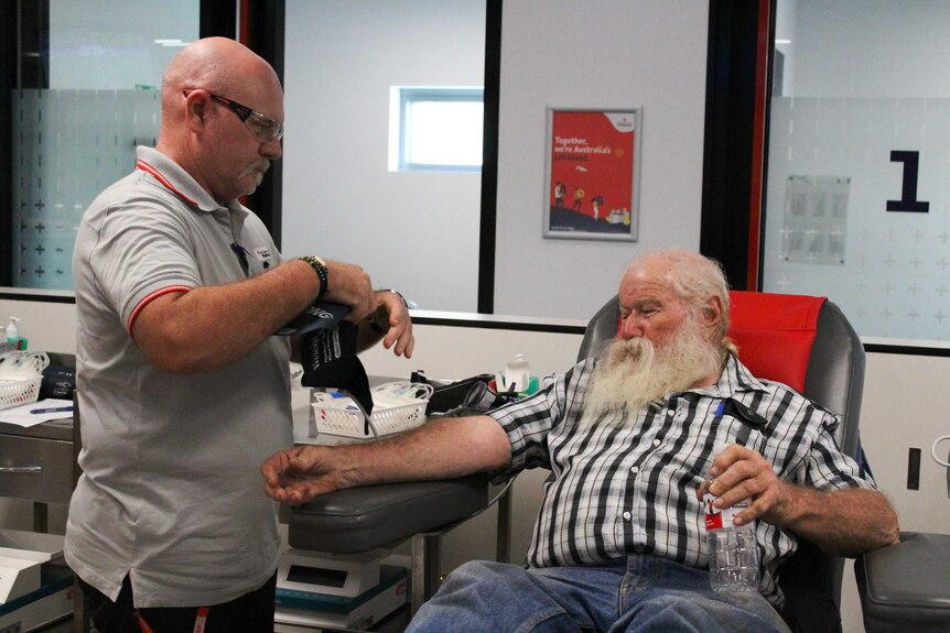 A man with a big white beard sits in a blood donation chair while a nurse prepares blood donation equipment to put on him