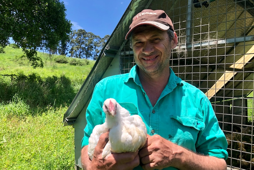 A man holding a chicken in a lush green pasture in front of a shed.
