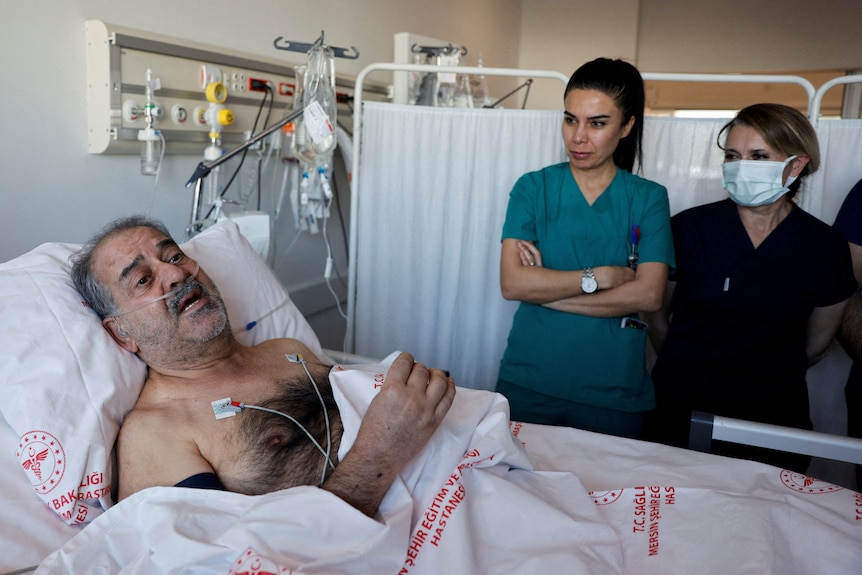 A man with greying stubble lies in a hospital bed with female medical staff standing next to him.