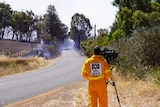 Cameraman wearing ABC News fire gear filming smoke in distance on country road.