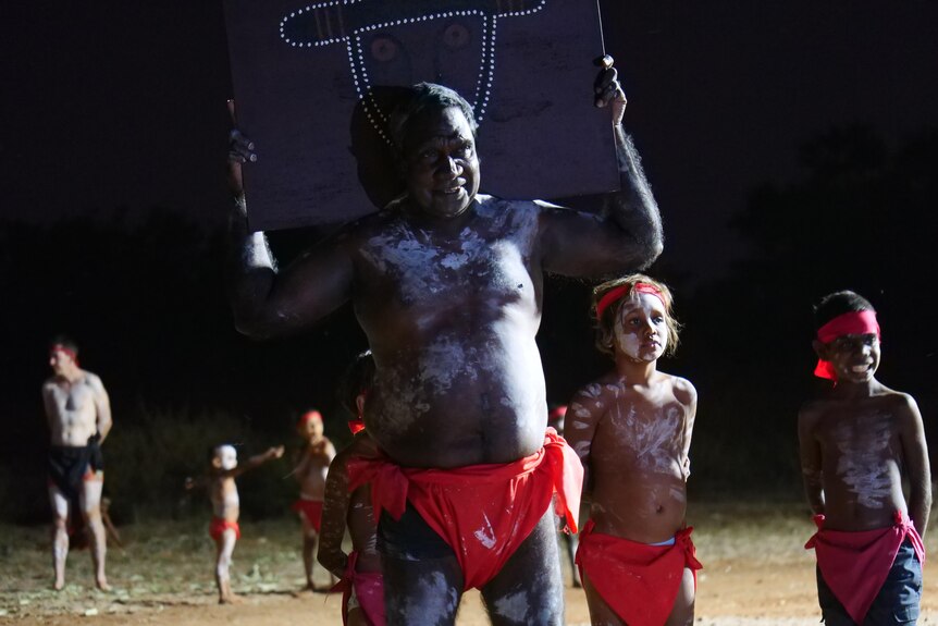 an Indigenous man, painted up, dances holding a wooden art work