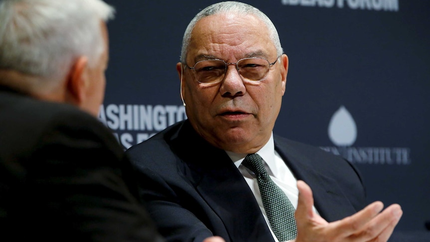 Two men with grey hair and wearing dark suits sit on a stage for a discussion in front of signage.