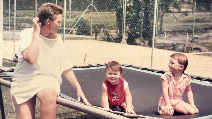 An old coloured profile of a smiling short-haired woman in short white dress sitting on a trampoline, with two happy children.