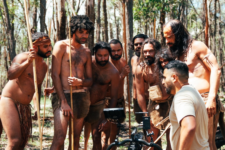 A group of Indigenous men in traditional dress holding spears look at a small camera screen.