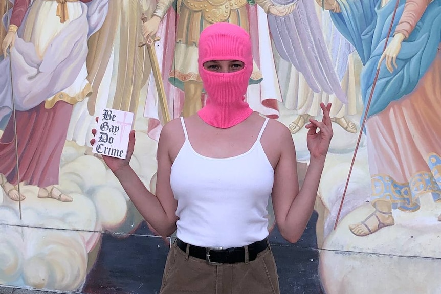 A queer Ukrainian activist stands in front of a religious scene in a pink balaclava holding a sign saying "Be gay, do crime". 