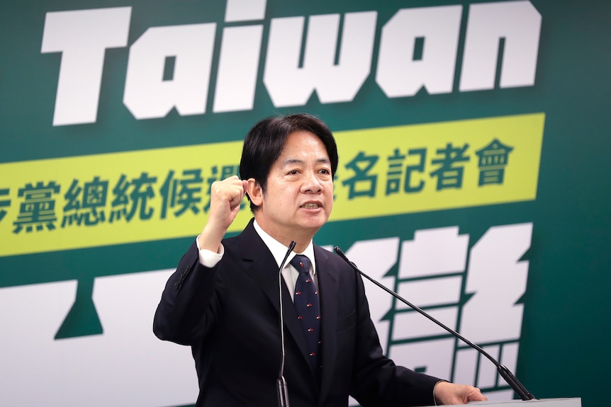 A man wearing a black suit with navy tie and red spots raises a fist in front of a sign that says Taiwan