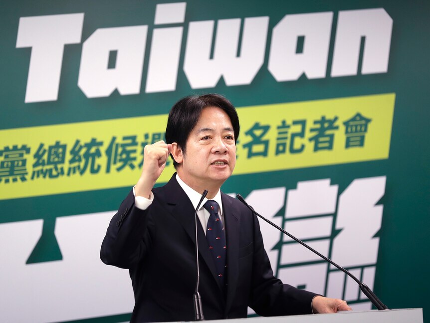 A man wearing a black suit with navy tie and red spots raises a fist in front of a sign that says Taiwan