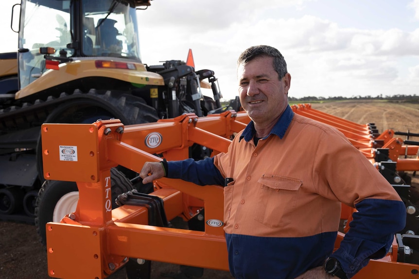 A smiling middle-aged man wearing an orange hi visibility shirt leans against an orange tractor.