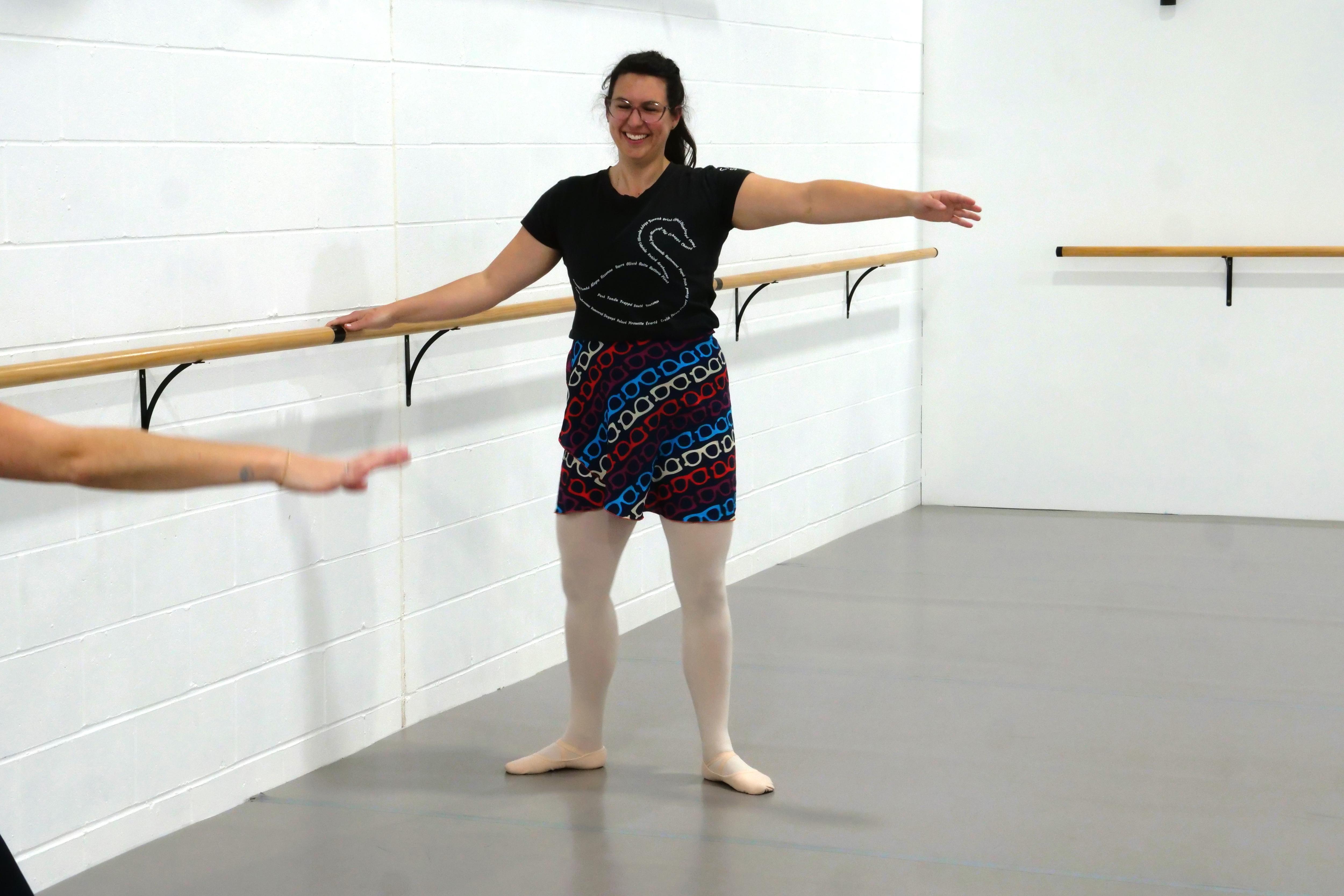 Kate holding the bar doing a ballet move with her arm outstretched. 