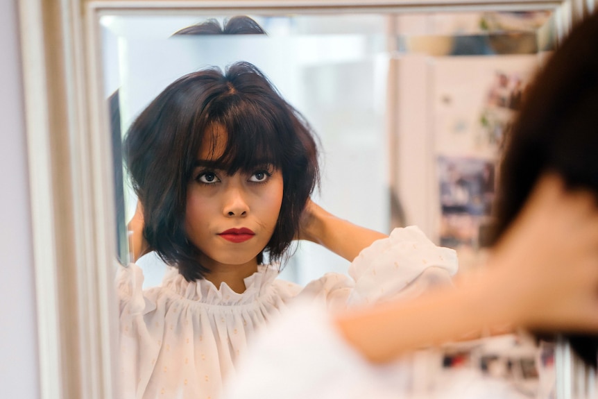 Woman with dark hair and red lipstick looks at herself in a mirror with a serious look on her face to depict bad body image