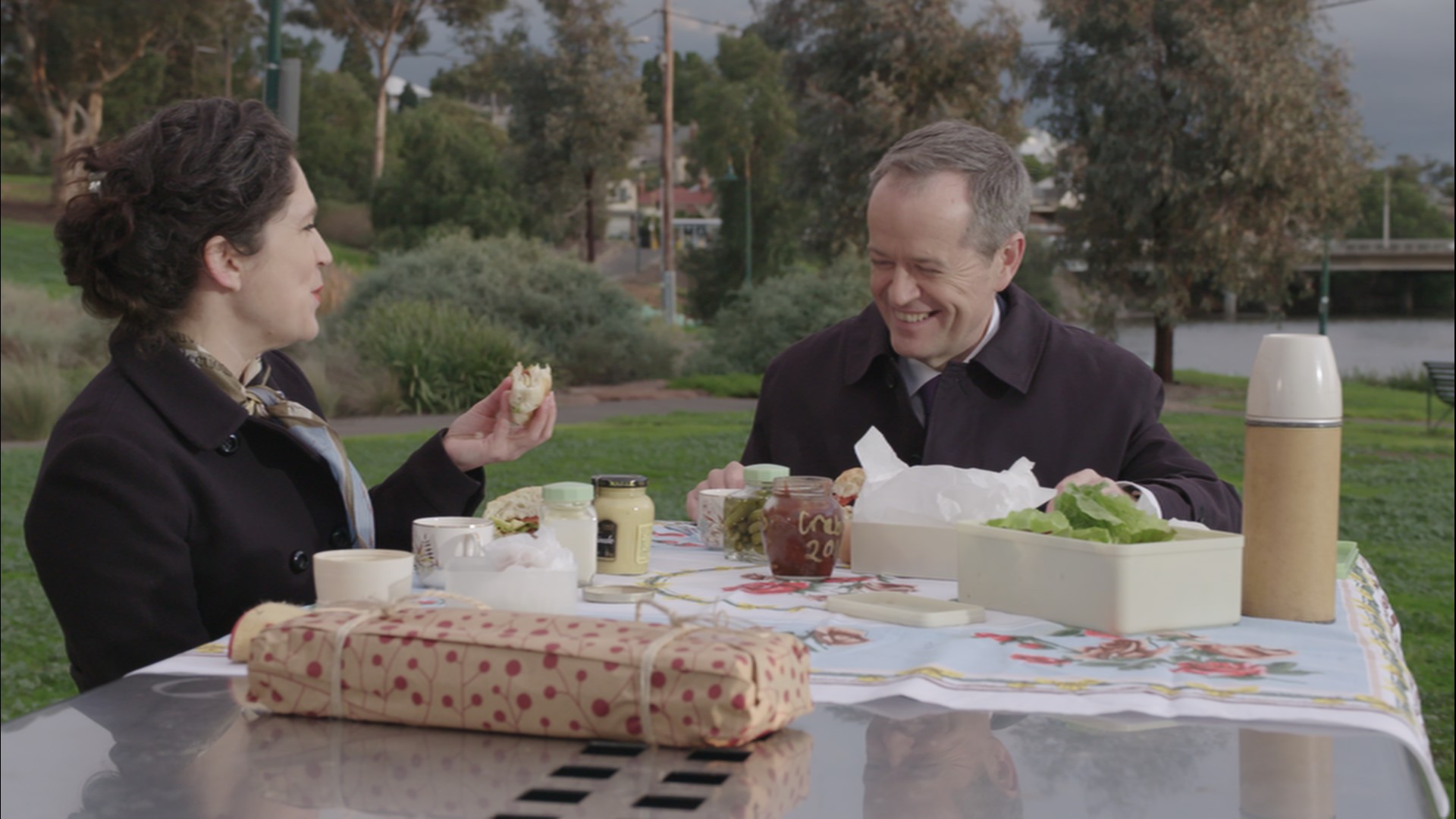 Woman and man sitting at picnic table in a park eating sandwiches and laughing.