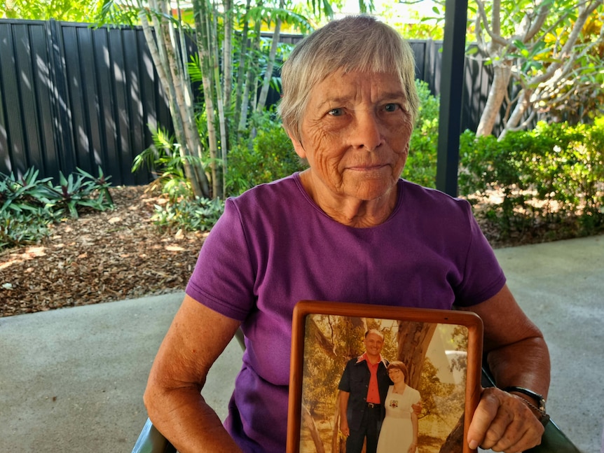 Judy sits in a chair looking serious, holding a photo of herself and her husband Bob from some decades earlier.