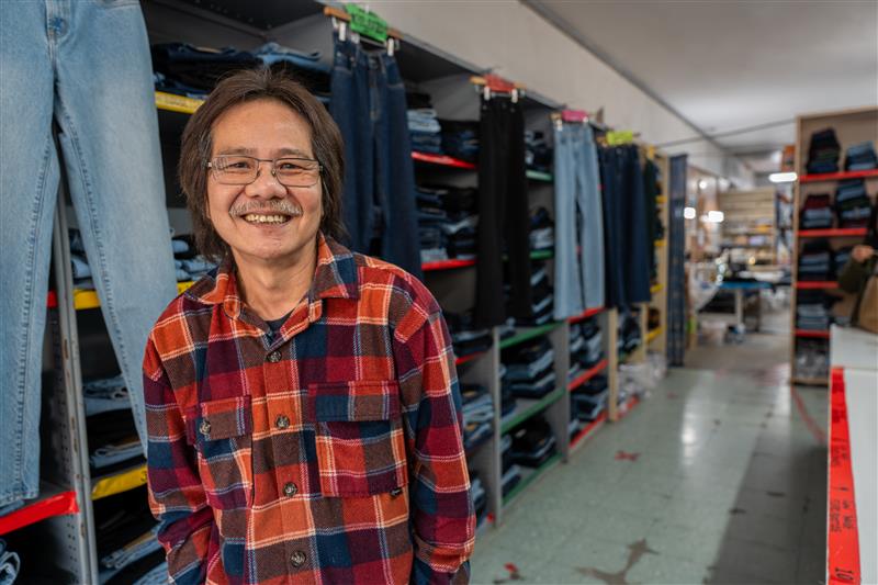 Man in check shirt and glasses smiles in front of racks of denim jeans