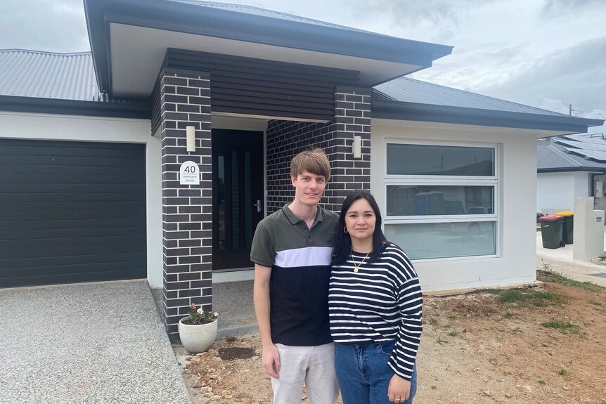 A young couple stand in front of a new house. 