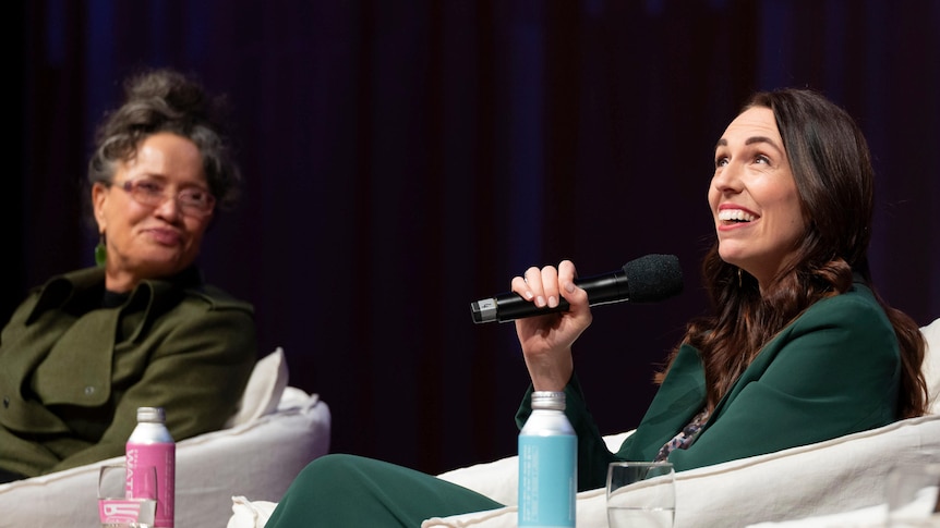 You view Jacinda Ardern in a green suit on a panel staring up while speaking into a microphone.