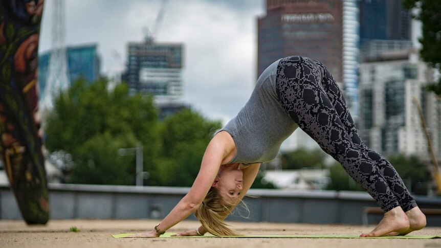 A young woman performs the downward facing dog pose on a yoga mat in an outdoor space.