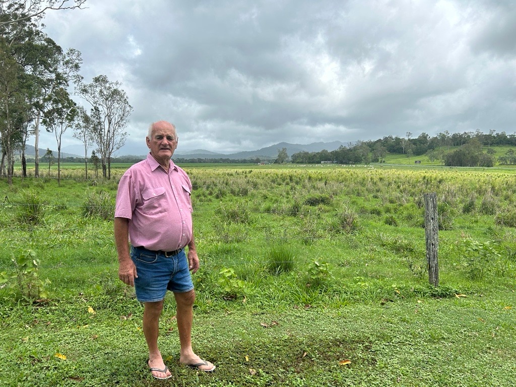 Bill Camm stands in front of a barb wire fence with a green paddock in the background and grey clouds.