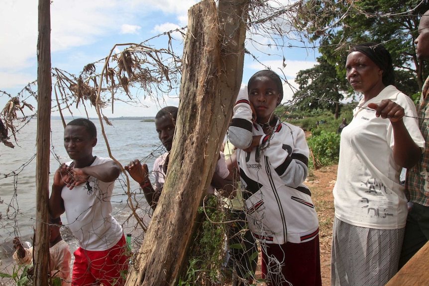 Two men and two women look through a damaged barbed wire fence next to the water.