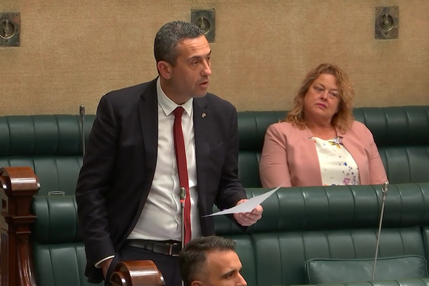 A man wearing a suit speaks while standing in Parliament while holding a piece of paper