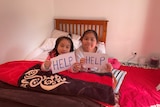 Two young girls on a bed with hand-written signs saying "HELP".