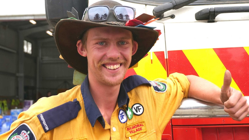 Man in bushman's hat and firefighter uniform grins and gives thumbs up as he leans on fire truck.