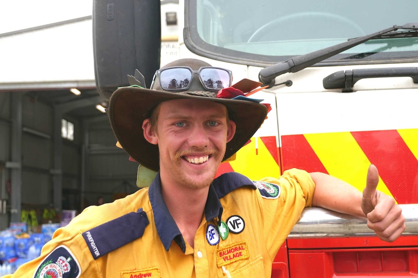 Man in bushman's hat and firefighter uniform grins and gives thumbs up as he leans on fire truck.